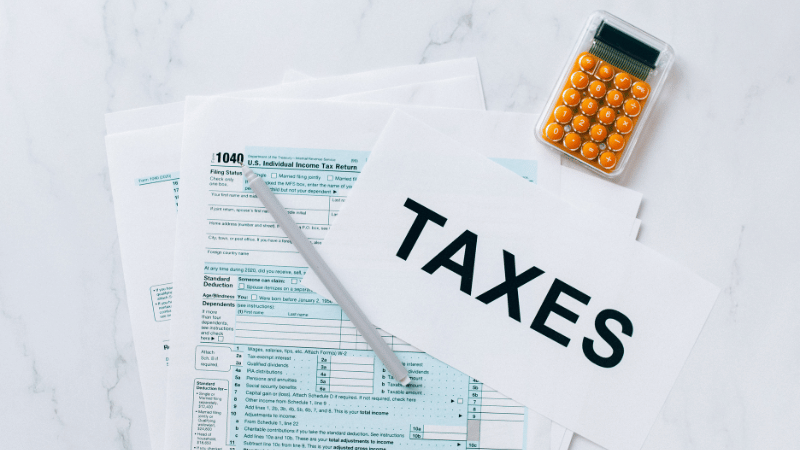 A pen and a calculator rest on top of U.S. Individual Income Tax Return forms, with a "TAXES" sign prominently displayed.