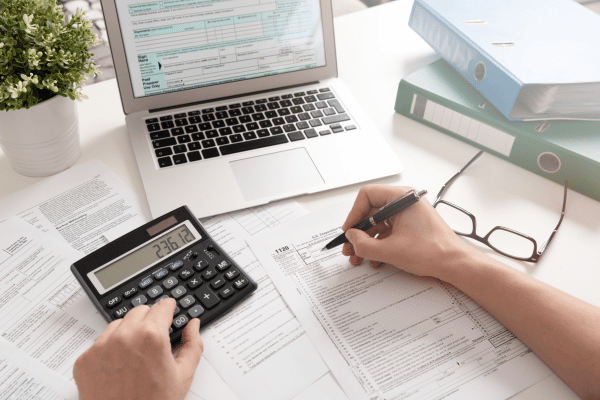 A person calculating taxes with a calculator, laptop, and various financial documents spread on the desk.